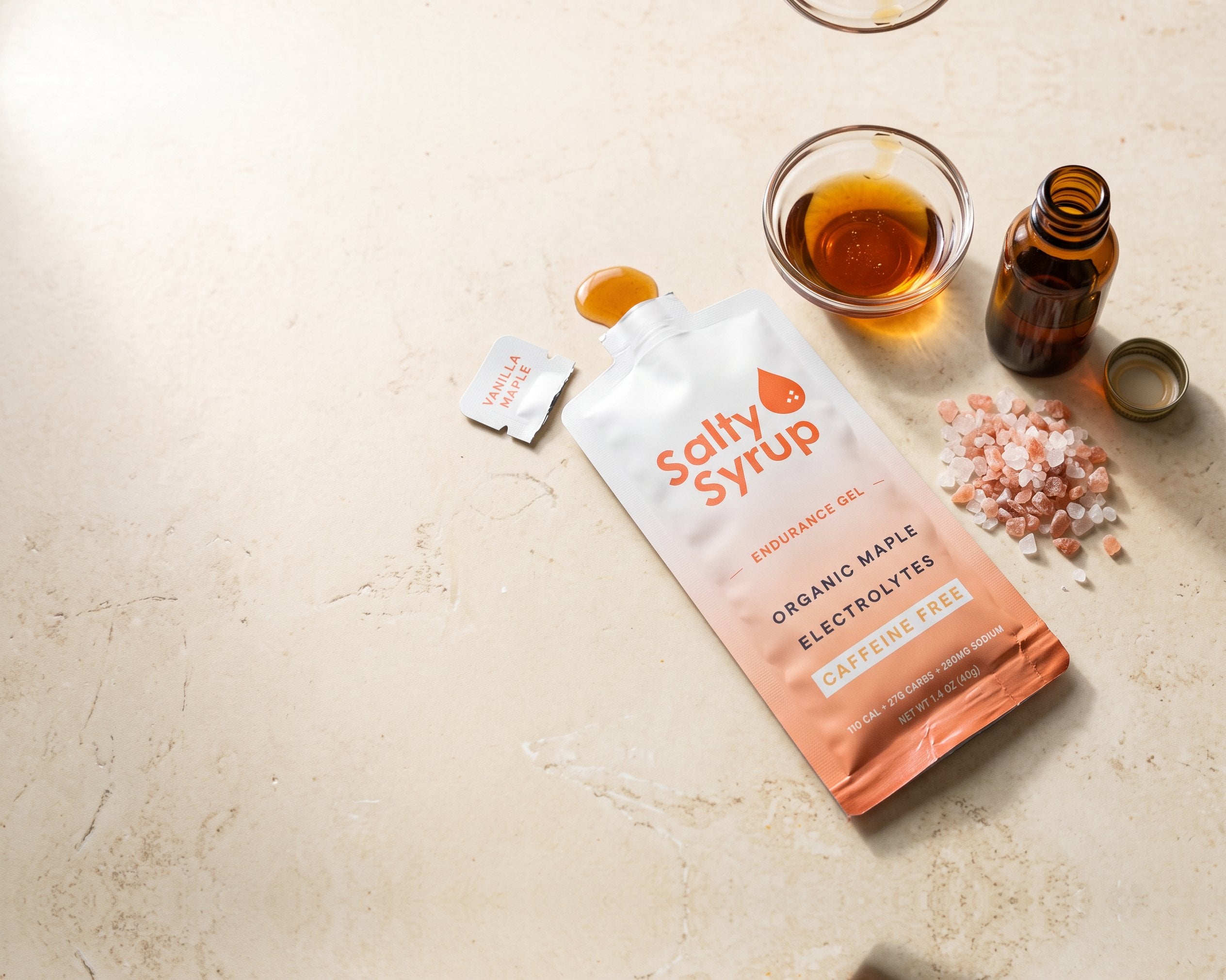 An opened Salty Syrup Vanilla Maple packet on stone beside its three ingredients: a dish of maple syrup, pink salt and a bottle of vanilla extract